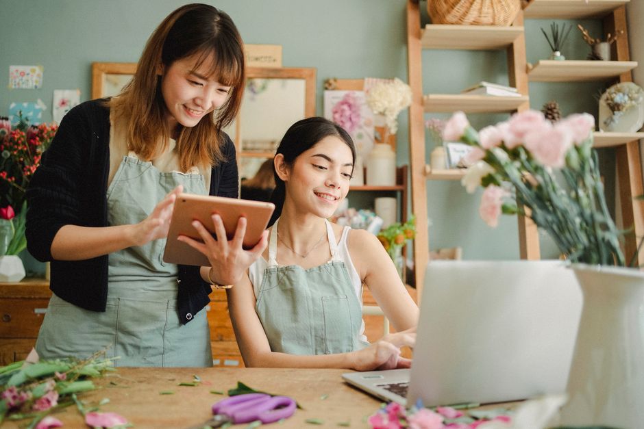 In a flower shop, two women wearing light green aprons work together on floral arrangements. One woman with shoulder-length brown hair, dressed in a black cardigan over a light top, is smiling while h