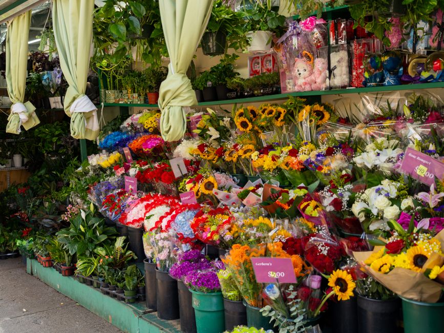 A vibrant flower stall displays an array of fresh floral arrangements and bouquets arranged on tiered shelves and in large buckets. The flowers include bright sunflowers, roses, lilies, and chrysanthe