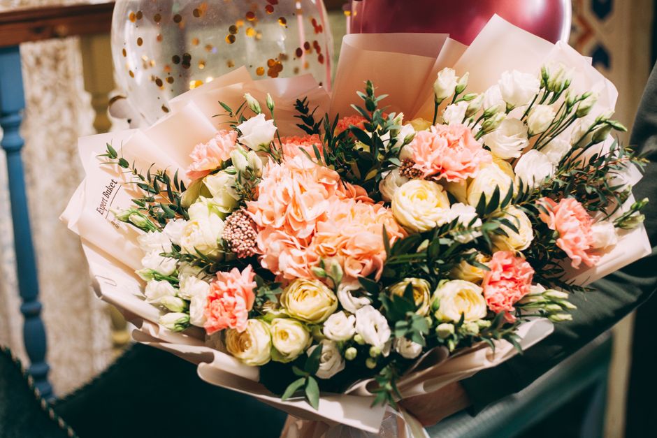 A vibrant floral bouquet featuring soft pink carnations, creamy white roses, and delicate white lisianthus, accented with lush green foliage. The flowers are arranged in a rounded, hand-tied style and