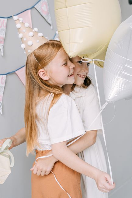 Two young girls celebrating a birthday indoors, with one wearing a light pink party hat decorated with white pom-poms. They are smiling and embracing each other while holding white and beige balloons,