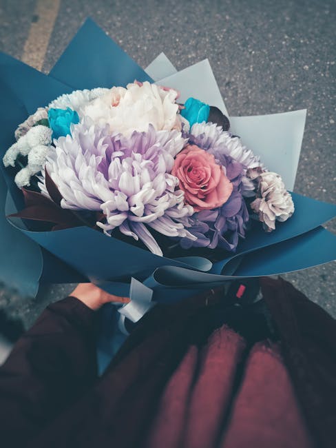 A vibrant floral bouquet featuring large white chrysanthemums, soft pink roses, and purple lisianthus, wrapped in blue and white paper with a matching ribbon. The arrangement is held in a person's han