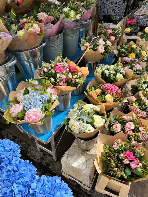 A vibrant display of freshly arranged floral bouquets and individual flower arrangements at a flower market. The bouquets feature pastel pink roses, white and pale yellow spray roses, light blue hydra