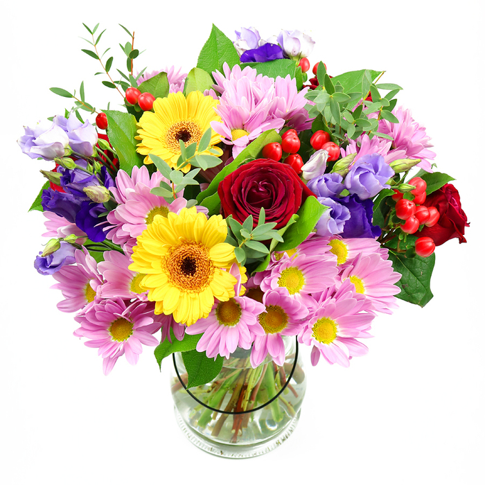 The photograph shows a vibrant, hand-crafted bouquet standing tall in a transparent glass vase, set against a pure white background that highlights every petal and leaf. At its centre, rich crimson red roses with velvety, tightly layered petals create a sense of depth and classic elegance, like the kind of timeless gift often chosen to celebrate anniversaries or special milestones in Barking. Flanking the roses, two radiant yellow gerbera daisies with dark, warm brown-orange centres radiate cheerful energy, their flat, daisy-like faces conveying an open, welcoming mood that would feel at home in a bright Barking living room or office near London Road. Surrounding these focal flowers, soft pink daisies or chrysanthemums fill the bouquet with volume; their many slender petals and small yellow centres create a gentle, almost feathery effect that softens the bolder tones. Interspersed throughout are graceful purple and lilac lisianthus blooms, their bell-shaped flowers and curled edges adding cooler notes and a refined, almost romantic lightness. Nestled between petals, clusters of glossy, round red hypericum berries offer a playful contrast in texture and an extra splash of colour. A mixture of greenery in various shades, from deep green broad leaves to lighter, more delicate sprigs, weaves between the flowers and extends beyond the rim of the vase, giving the arrangement a full, flourishing silhouette. Through the clear glass, neatly cut stems fan out in clean water, showing the professional care that went into conditioning each stem. Crafted with the attention to detail you'd expect from a dedicated Barking florist, this bouquet feels ideal for birthdays, thank-you gestures, or simply bringing a burst of fresh, colourful joy into a home.